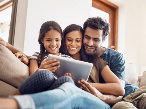 A family of three smiling and looking at a tablet together on a couch.