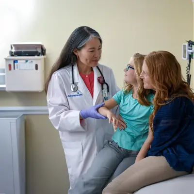 AdventHealth Centra Care physician in white coat examining a young girl's arm while her mother watches during a consultation in a clinic room