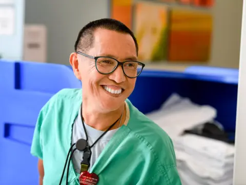 A smiling healthcare worker in green scrubs stands in a hospital room, exuding warmth and positivity.
