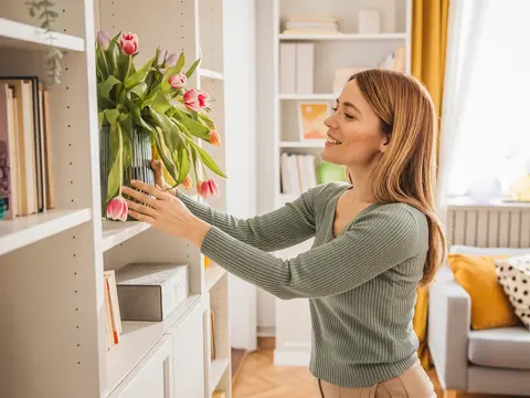 Smiling woman in a green striped sweater placing a vase of pink tulips on a white bookshelf in a bright, modern living room with yellow curtains and a cozy sofa in the background