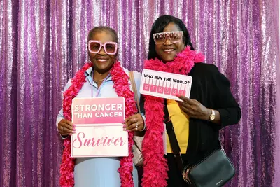 Two women wearing pink boas and festive glasses hold "Stronger Than Cancer Survivor" and "Who Run the World? Girls!" signs in front of a purple sequin backdrop.