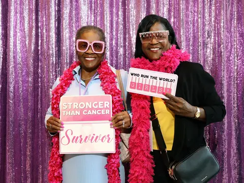 Two women wearing pink boas and festive glasses hold "Stronger Than Cancer Survivor" and "Who Run the World? Girls!" signs in front of a purple sequin backdrop.
