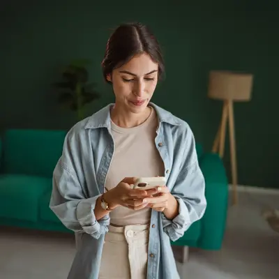 Woman in light blue denim shirt looking down at her phone while standing in a modern living room with a green couch