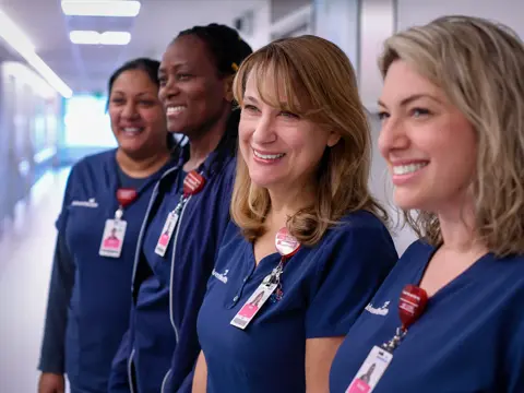 Four smiling AdventHealth nurses in blue scrubs stand together in a hospital hallway.