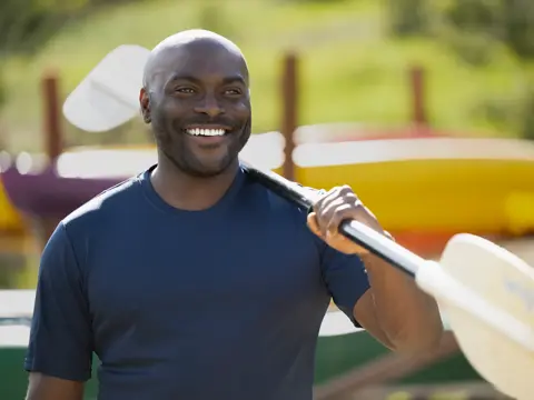 A smiling man holding a paddle in front of colorful kayaks in a park.
