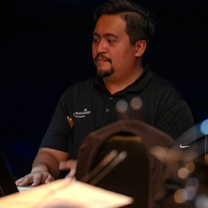 Jerel Moral performs with the orchestra, wearing an AdventHealth polo shirt, standing behind percussion instruments with mallets in motion during a performance