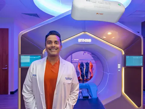 Doctor in white coat and orange scrubs smiling in front of an advanced radiation therapy machine in a modern treatment room with purple ambient lighting