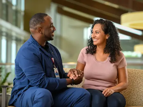 A smiling man and woman sit together on a couch, holding hands and conversing.