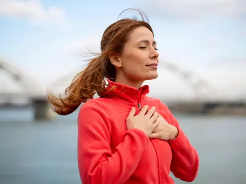 Person in a bright pink fleece stands by the water with eyes closed and hands on their chest, with a bridge in the background.
