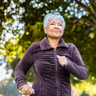 Older woman in purple jacket jogging outdoors in a park with trees in the background