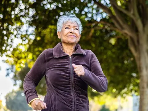 Older woman in purple jacket jogging outdoors in a park with trees in the background