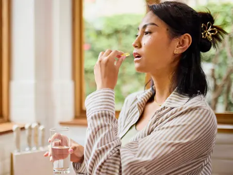 Woman in striped robe taking a supplement pill with a glass of water in a bright home setting near a window