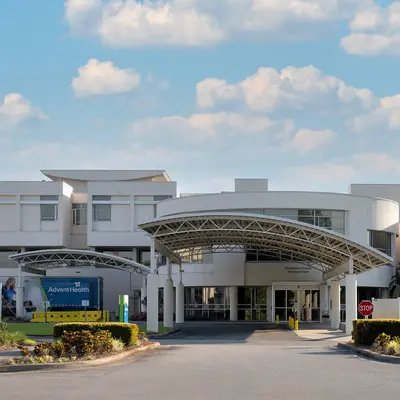 Modern white hospital building entrance with curved metal canopy, palm trees on either side, and AdventHealth signage visible at the covered entry drive