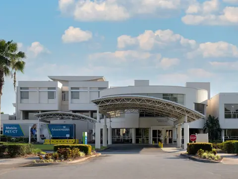 Modern white hospital building entrance with curved metal canopy, palm trees on either side, and AdventHealth signage visible at the covered entry drive