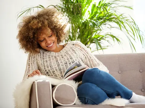 A woman smiling while sitting on the couch reading.