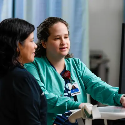 Two health care team members in teal scrubs sitting at a desk, looking at a computer monitor together in a clinical setting with blue privacy curtains in the background.