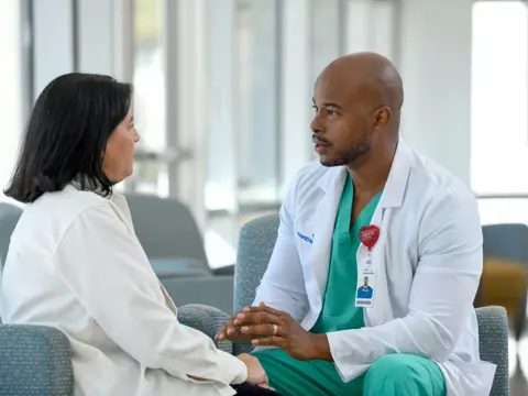A male doctor in a white coat and green scrubs talks with a female patient in a hospital waiting room.