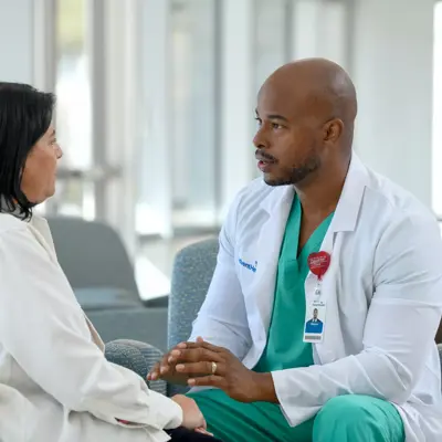 A male doctor in a white coat and green scrubs talks with a female patient in a hospital waiting room.