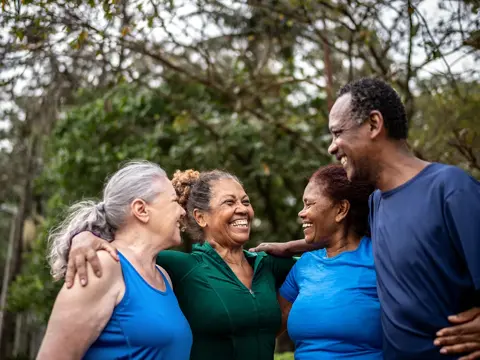 Four older adults in athletic clothing smiling together with their arms around one another outdoors.