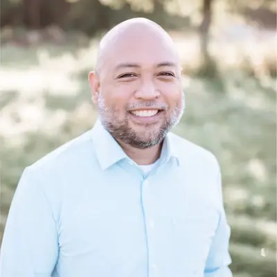 A smiling man with a bald head and gray beard wearing a light blue button-down shirt, photographed outdoors with a soft-focus natural background