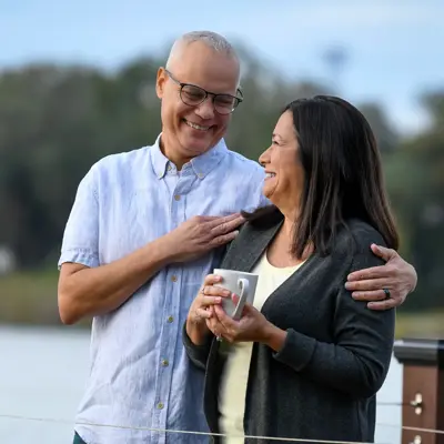 Older couple smiling and embracing on a wooden deck overlooking a lake, with the woman holding a white mug