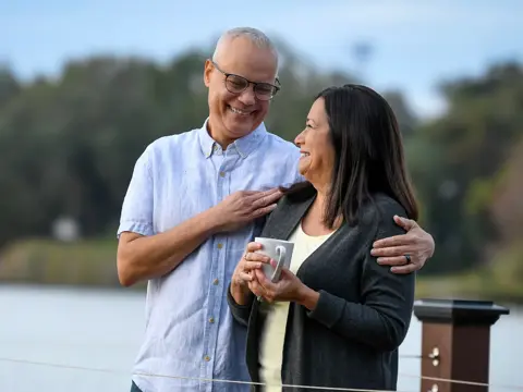 Older couple smiling and embracing on a wooden deck overlooking a lake, with the woman holding a white mug