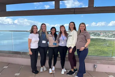 Six medical residents in casual attire stand together on an outdoor terrace, smiling at the camera with a waterfront and city skyline in the background