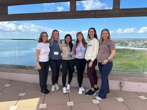 Six medical residents in casual attire stand together on an outdoor terrace, smiling at the camera with a waterfront and city skyline in the background