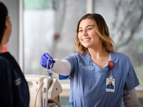 A smiling nurse in blue scrubs and a badge stands in a hospital corridor, holding a device.