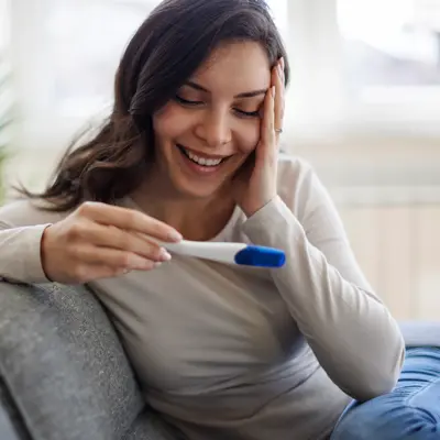 Smiling pregnant woman sitting on couch holding a positive pregnancy test with a blue indicator