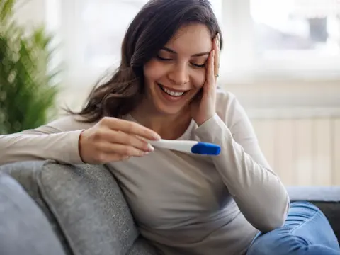 Smiling pregnant woman sitting on couch holding a positive pregnancy test with a blue indicator