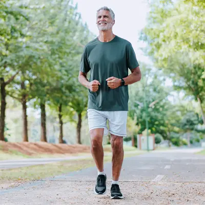 Older man jogging on a tree-lined path, wearing a dark shirt, light shorts, and earbuds.