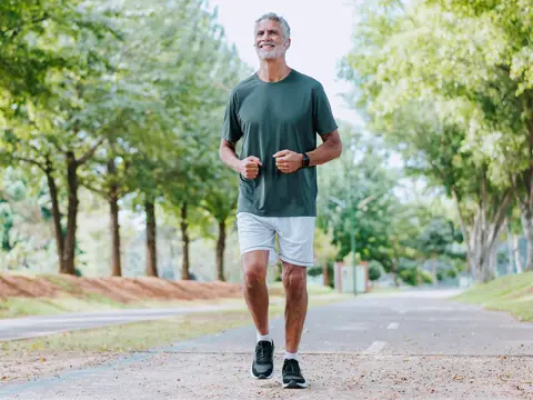 Older man jogging on a tree-lined path, wearing a dark shirt, light shorts, and earbuds.
