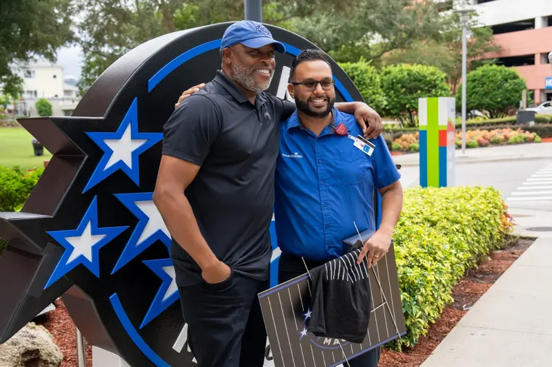 Two men standing in front of a big Orlando Magic logo outdoors.