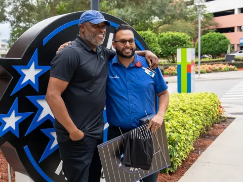 Two men standing in front of a big Orlando Magic logo outdoors.