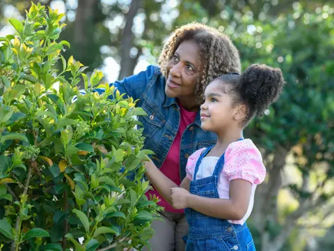 A smiling grandmother and granddaughter explore a bush together in a garden.