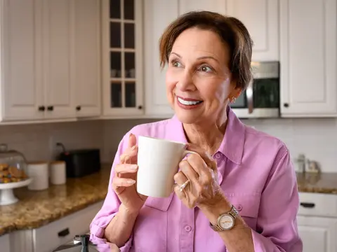 A smiling woman in a pink shirt holds a white mug in a modern kitchen.