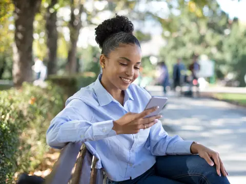A young woman in a light blue shirt sitting outdoors on a park bench, smiling while looking at her phone.
