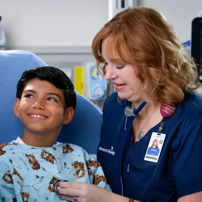 A smiling nurse checks a young patient's heart with a stethoscope in a hospital room.