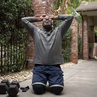 A man in workout clothes stretches on a driveway with dumbbells nearby, smiling.