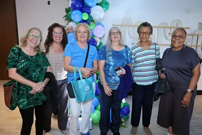 Six women smiling together in front of a blue and green balloon display at an event