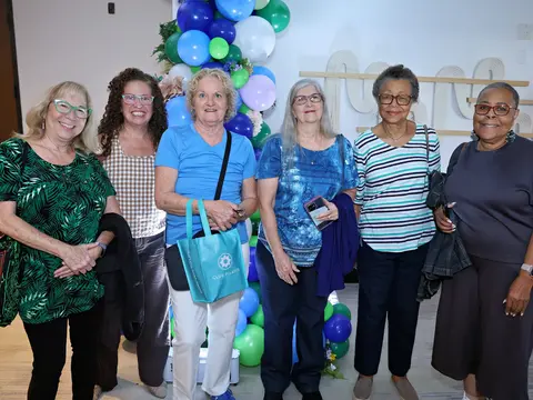 Six women smiling together in front of a blue and green balloon display at an event