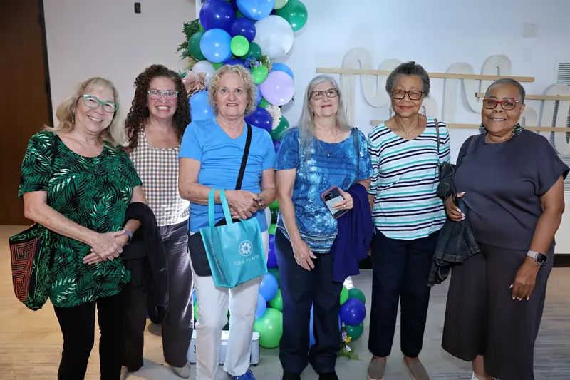 Six women smiling together in front of a blue and green balloon display at an event