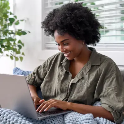 A woman with curly hair is sitting on a couch, smiling while using a laptop.