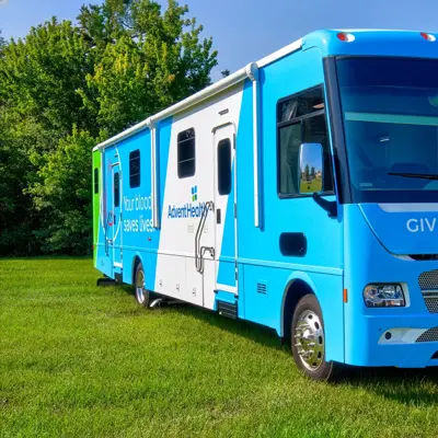 Blue and white AdventHealth bus on grass with text “Give Blood Today” and “Your blood saves lives.”