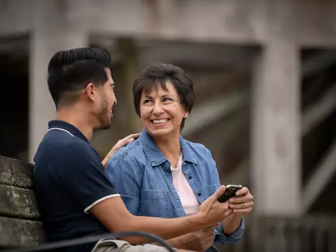A smiling woman and a man sit on a bench, sharing a moment while she looks at her phone.