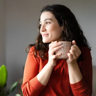 Young happy beautiful woman holding a cup of hot drink and looking trough the window feeling whole.