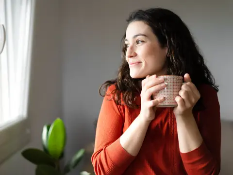 Young happy beautiful woman holding a cup of hot drink and looking trough the window feeling whole.