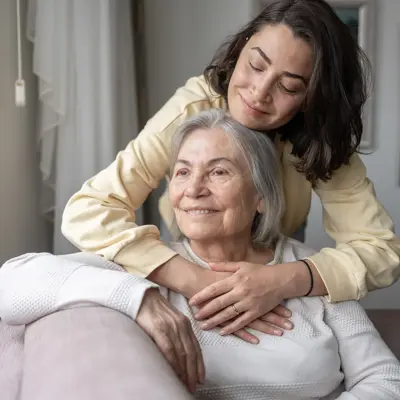 A middle-aged woman in a yellow sweater lovingly embraces her senior mother from behind as they sit together in a comfortable home setting