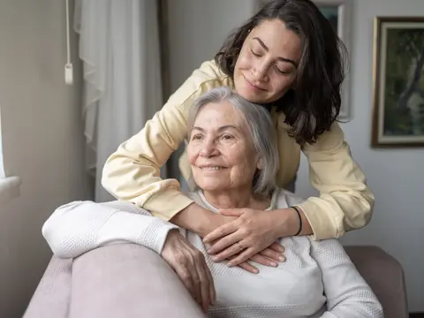 A middle-aged woman in a yellow sweater lovingly embraces her senior mother from behind as they sit together in a comfortable home setting